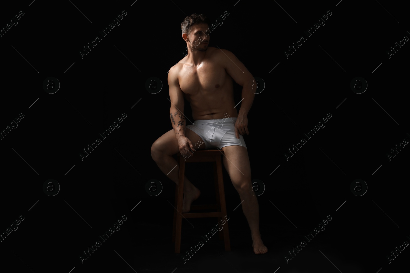 Young man in stylish white underwear on chair against black background Photo of Young man in stylish white underwear on chair against black background