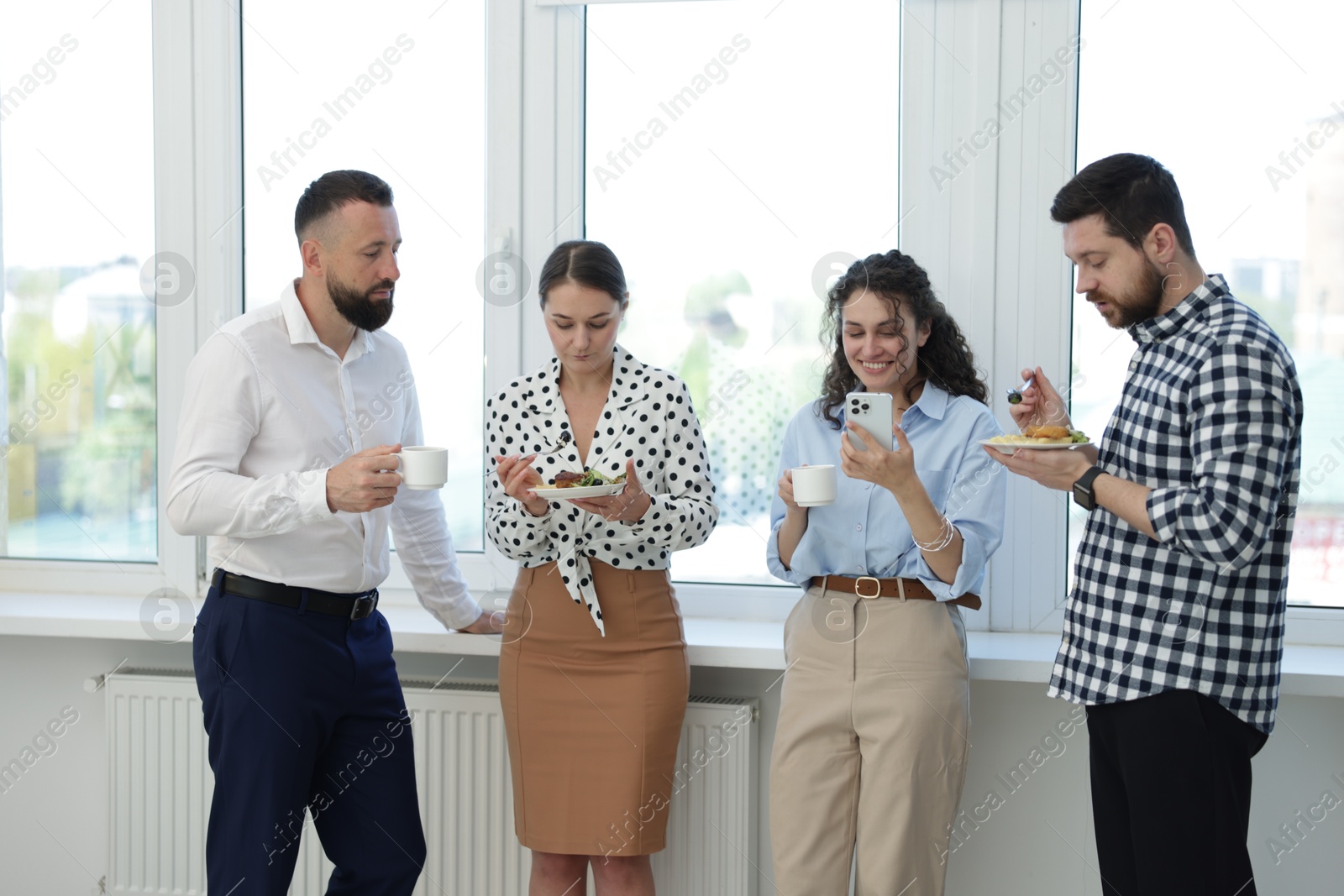 Colleagues with food and drinks near window in office during lunch break Photo of Colleagues with food and drinks near window in office during lunch break