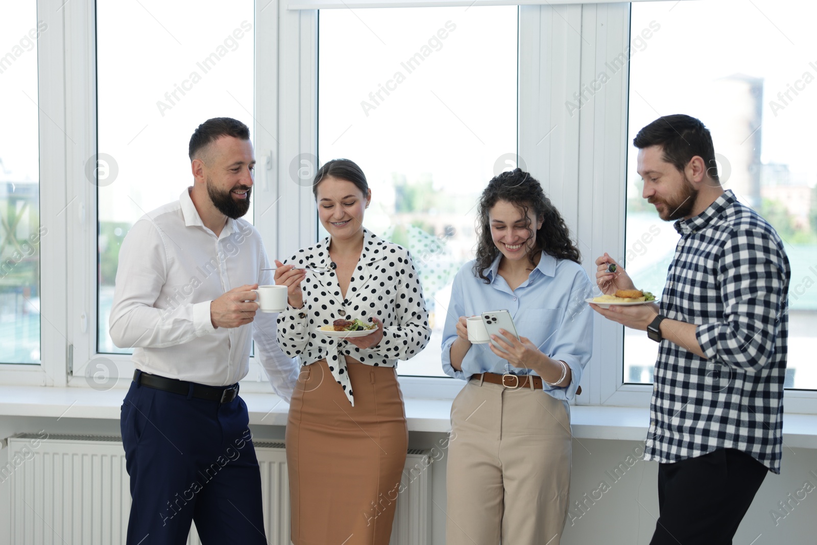 Colleagues with food and drinks near window in office during lunch break Photo of Colleagues with food and drinks near window in office during lunch break