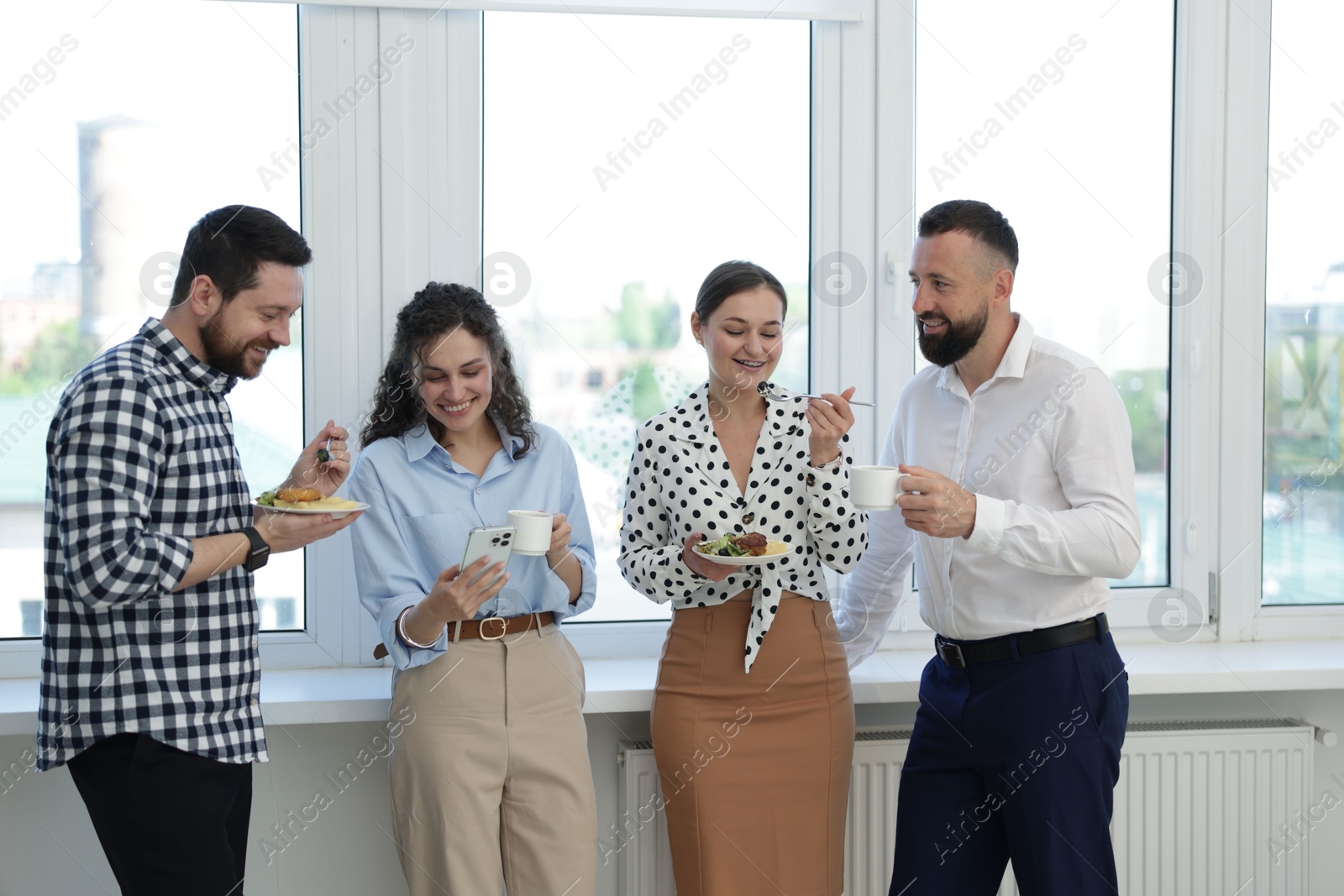 Colleagues with food and drinks near window in office during lunch break Photo of Colleagues with food and drinks near window in office during lunch break