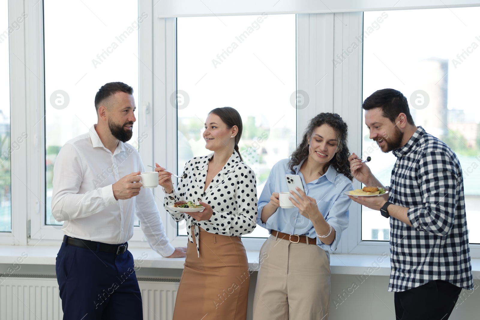 Colleagues with food and drinks near window in office during lunch break Photo of Colleagues with food and drinks near window in office during lunch break