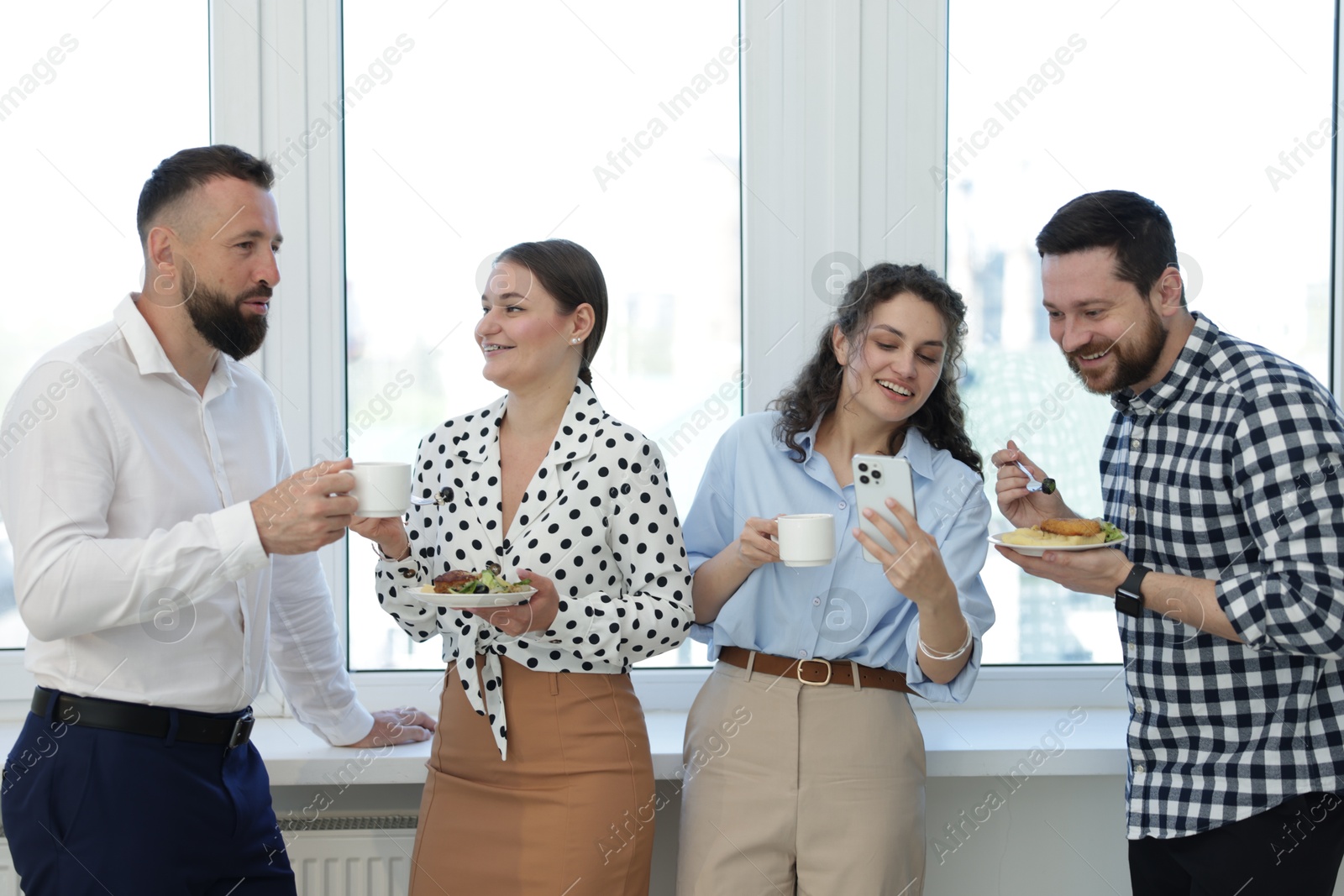 Colleagues with food and drinks near window in office during lunch break Photo of Colleagues with food and drinks near window in office during lunch break