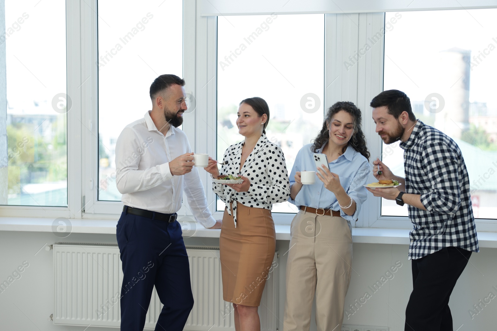 Colleagues with food and drinks near window in office during lunch break Photo of Colleagues with food and drinks near window in office during lunch break