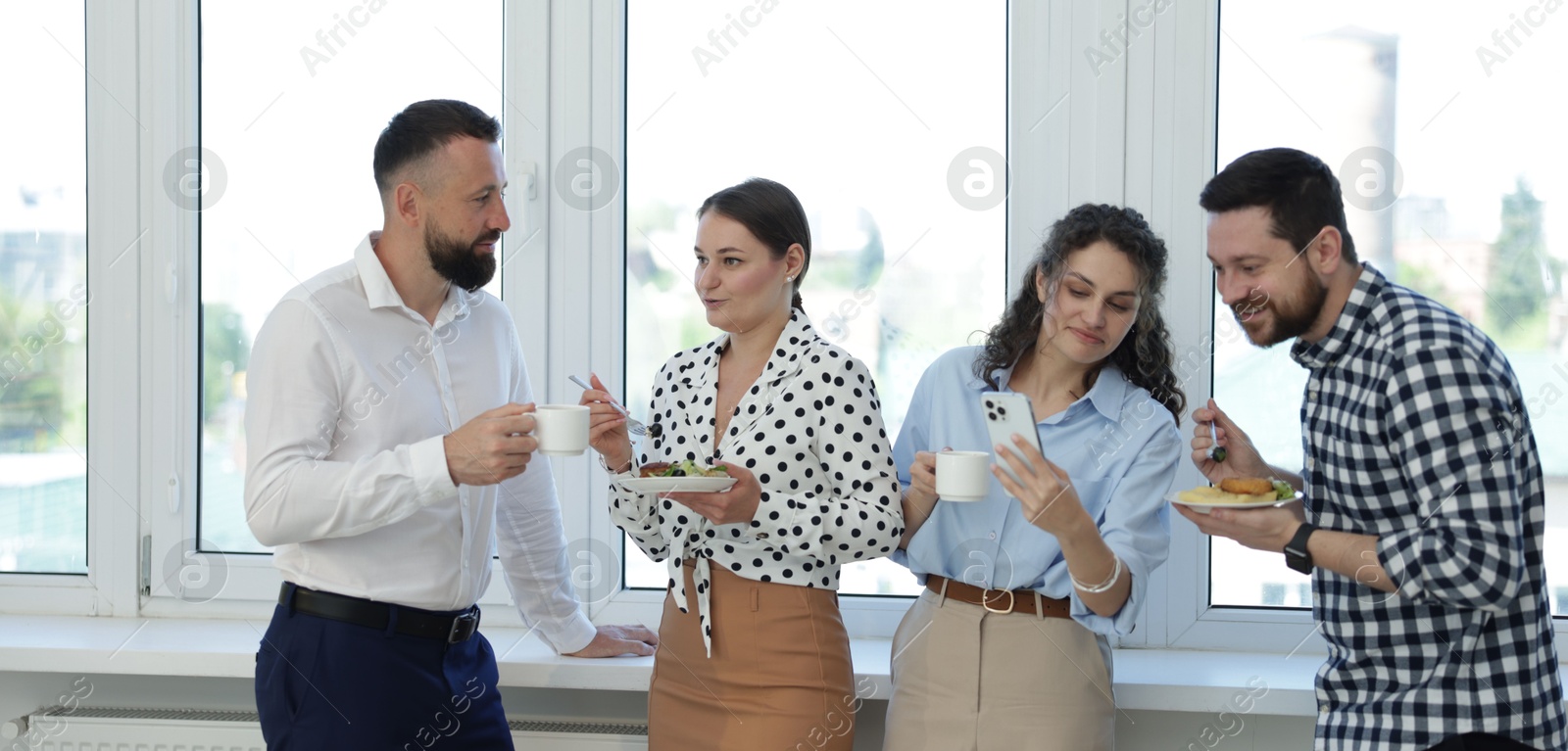 Colleagues with food and drinks near window in office during lunch break Photo of Colleagues with food and drinks near window in office during lunch break