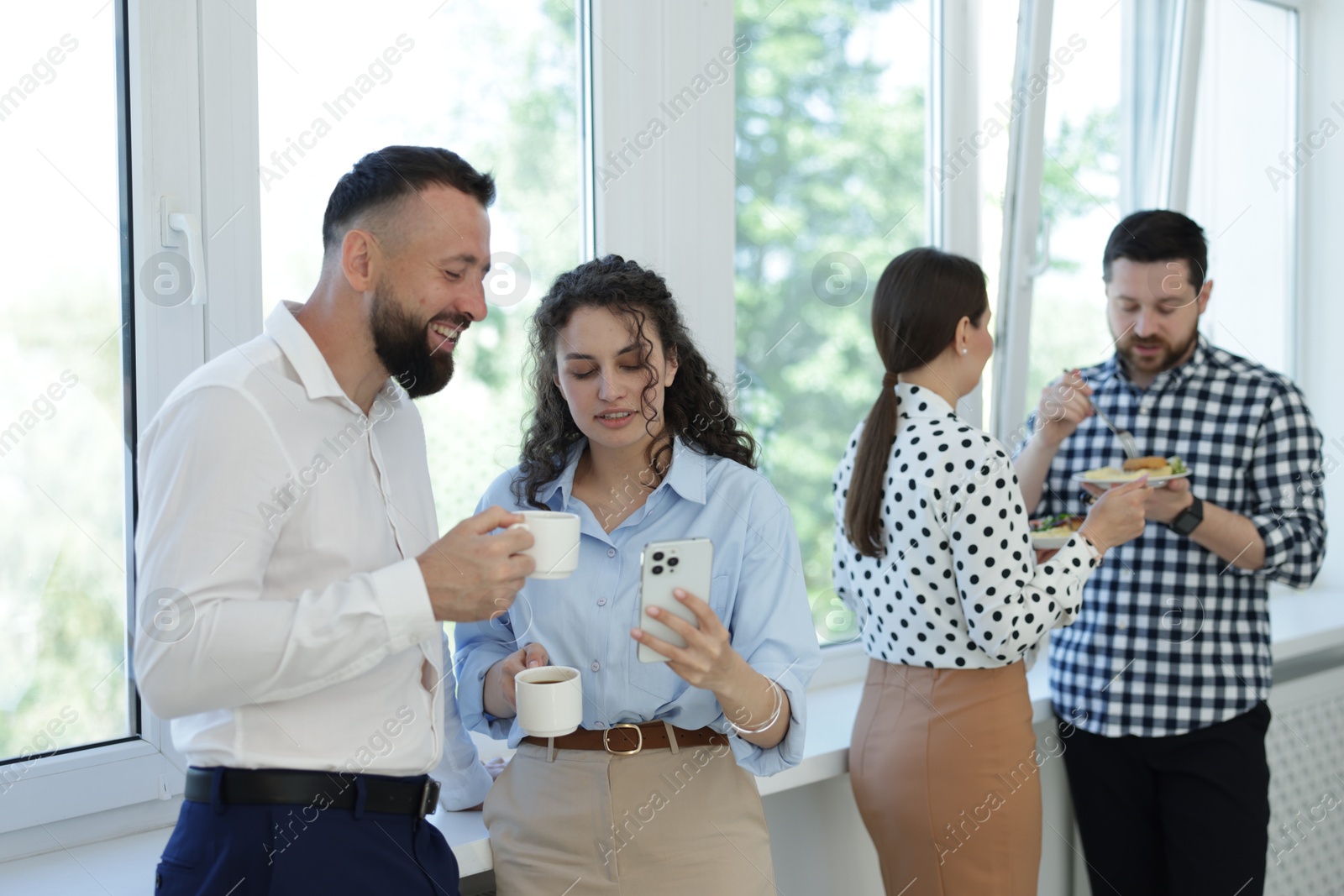 Colleagues with food and drinks near window in office during lunch break Photo of Colleagues with food and drinks near window in office during lunch break