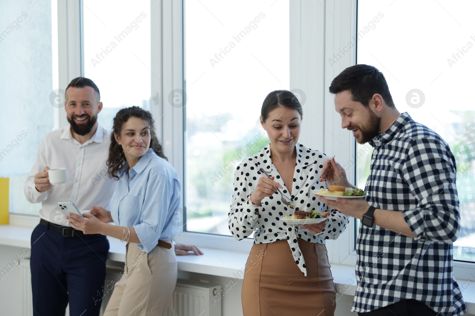 Colleagues with food and drinks near window in office during lunch break Photo of Colleagues with food and drinks near window in office during lunch break
