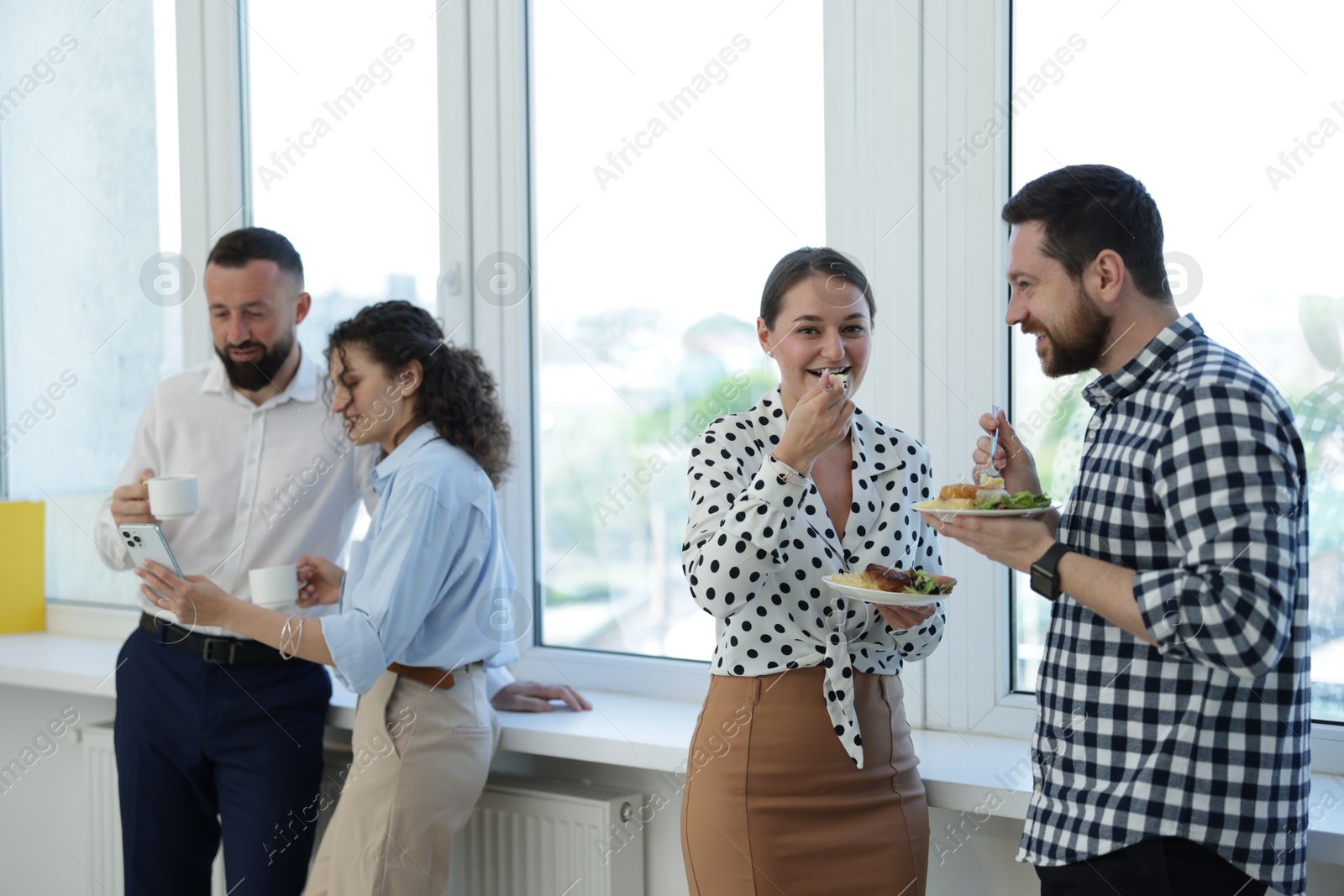 Colleagues with food and drinks near window in office during lunch break Photo of Colleagues with food and drinks near window in office during lunch break