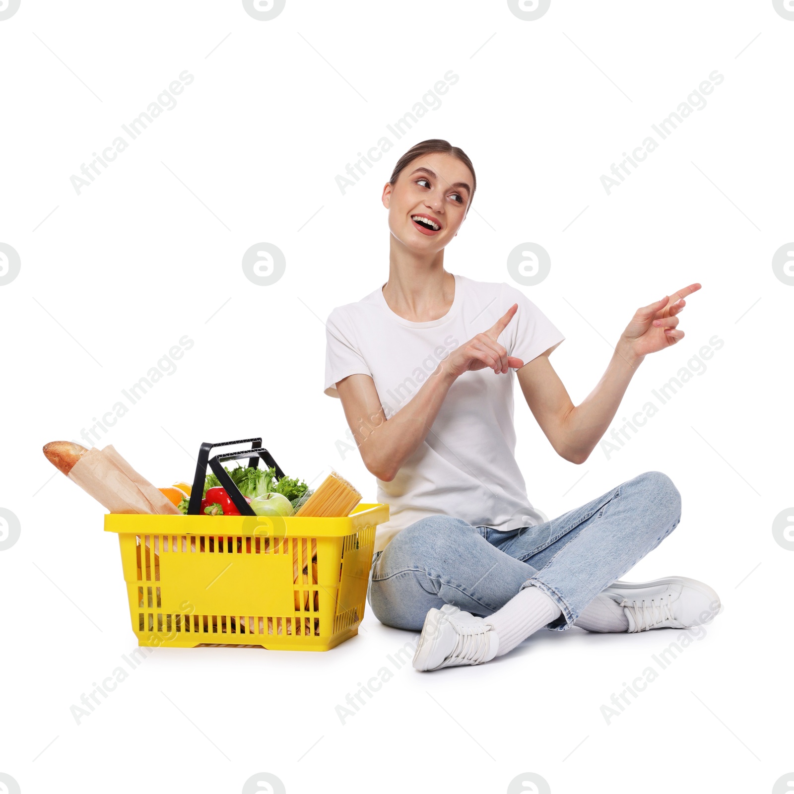 Woman with shopping basket full of products pointing at something on white background Photo of Woman with shopping basket full of products pointing at something on white background