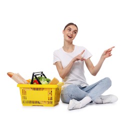 Photo of Woman with shopping basket full of products pointing at something on white background