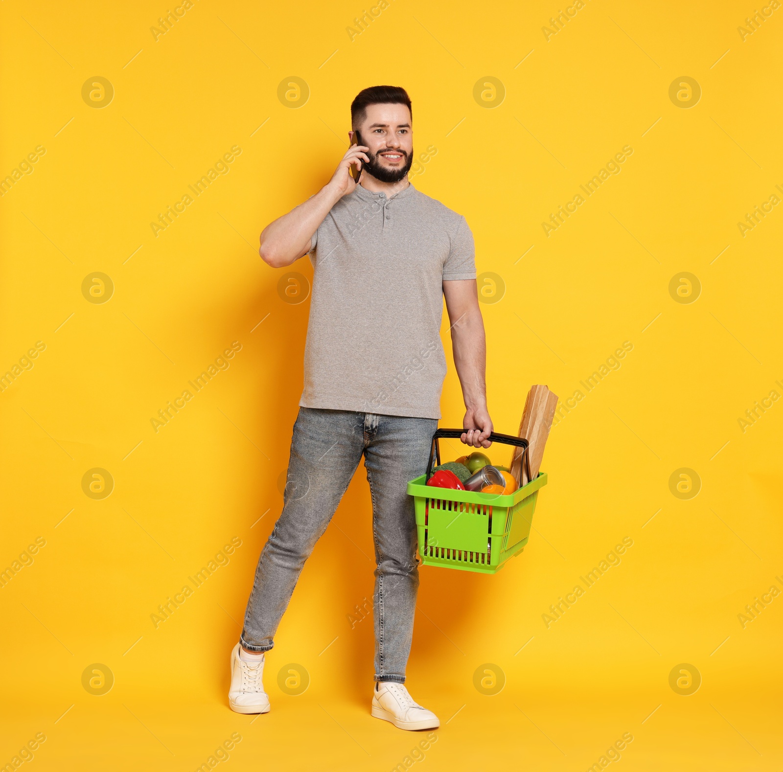 Man with shopping basket full of products talking on smartphone against orange background Photo of Man with shopping basket full of products talking on smartphone against orange background
