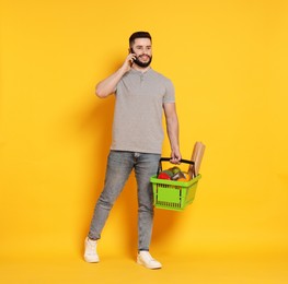 Photo of Man with shopping basket full of products talking on smartphone against orange background