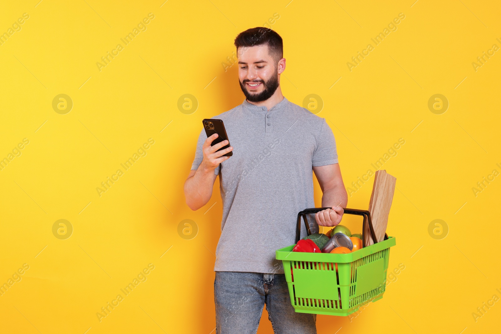 Man with shopping basket full of products and smartphone on orange background, space for text Photo of Man with shopping basket full of products and smartphone on orange background, space for text