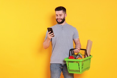 Photo of Man with shopping basket full of products and smartphone on orange background, space for text
