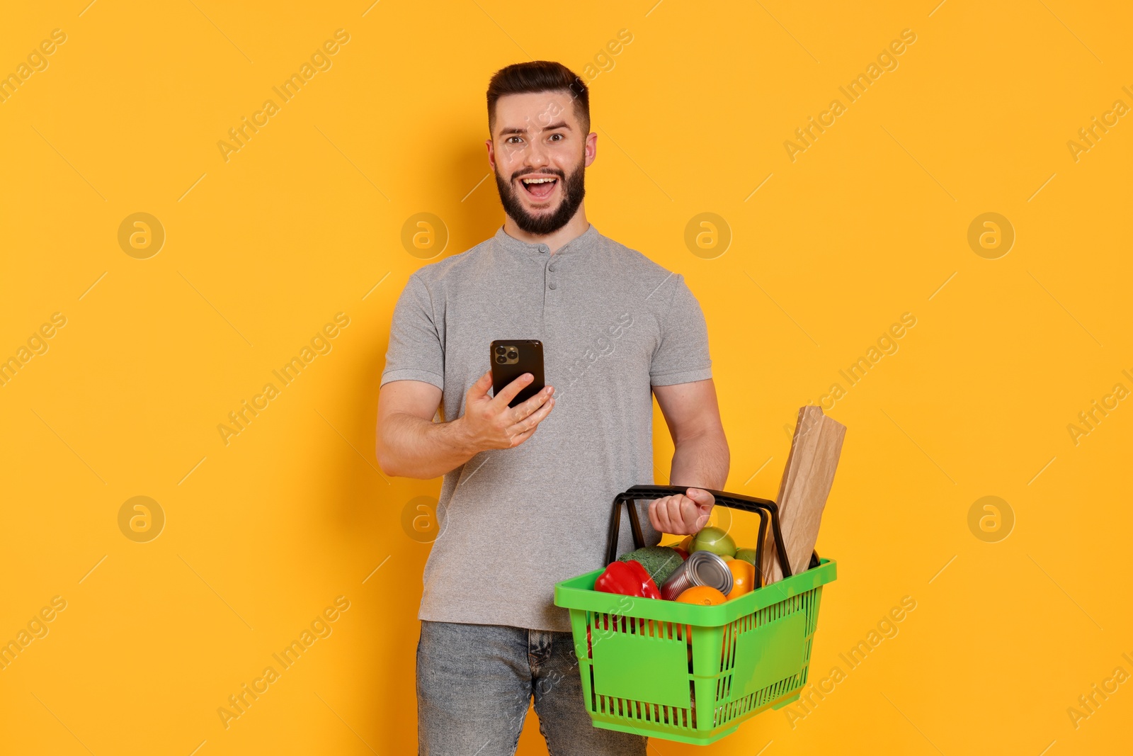 Man with shopping basket full of products and smartphone on orange background Photo of Man with shopping basket full of products and smartphone on orange background