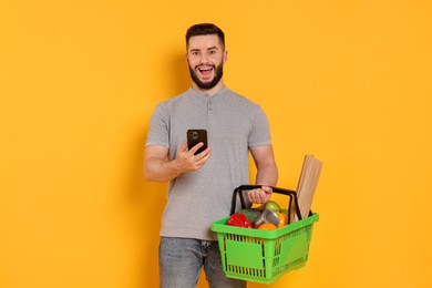 Photo of Man with shopping basket full of products and smartphone on orange background