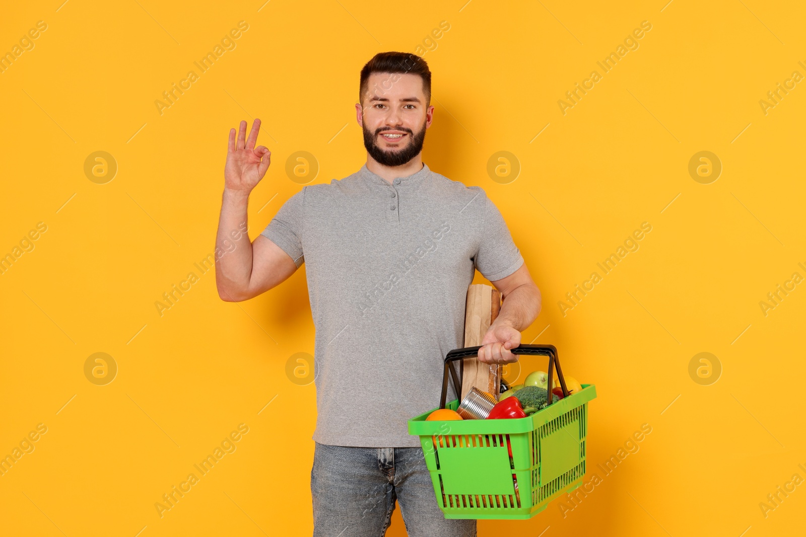 Man with shopping basket full of products showing ok gesture on orange background Photo of Man with shopping basket full of products showing ok gesture on orange background