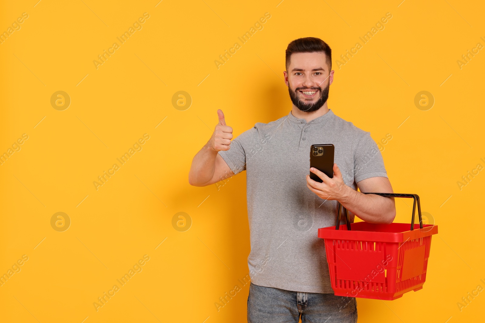 Man with empty shopping basket and smartphone showing thumbs up on orange background, space for text Photo of Man with empty shopping basket and smartphone showing thumbs up on orange background, space for text