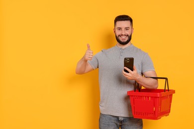 Photo of Man with empty shopping basket and smartphone showing thumbs up on orange background, space for text