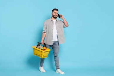 Photo of Man with shopping basket full of products talking on smartphone against light blue background