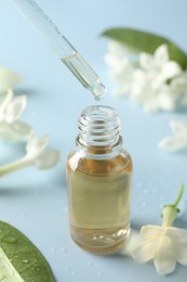 Photo of Dripping essential oil from pipette into bottle and jasmine flowers on light blue background, closeup