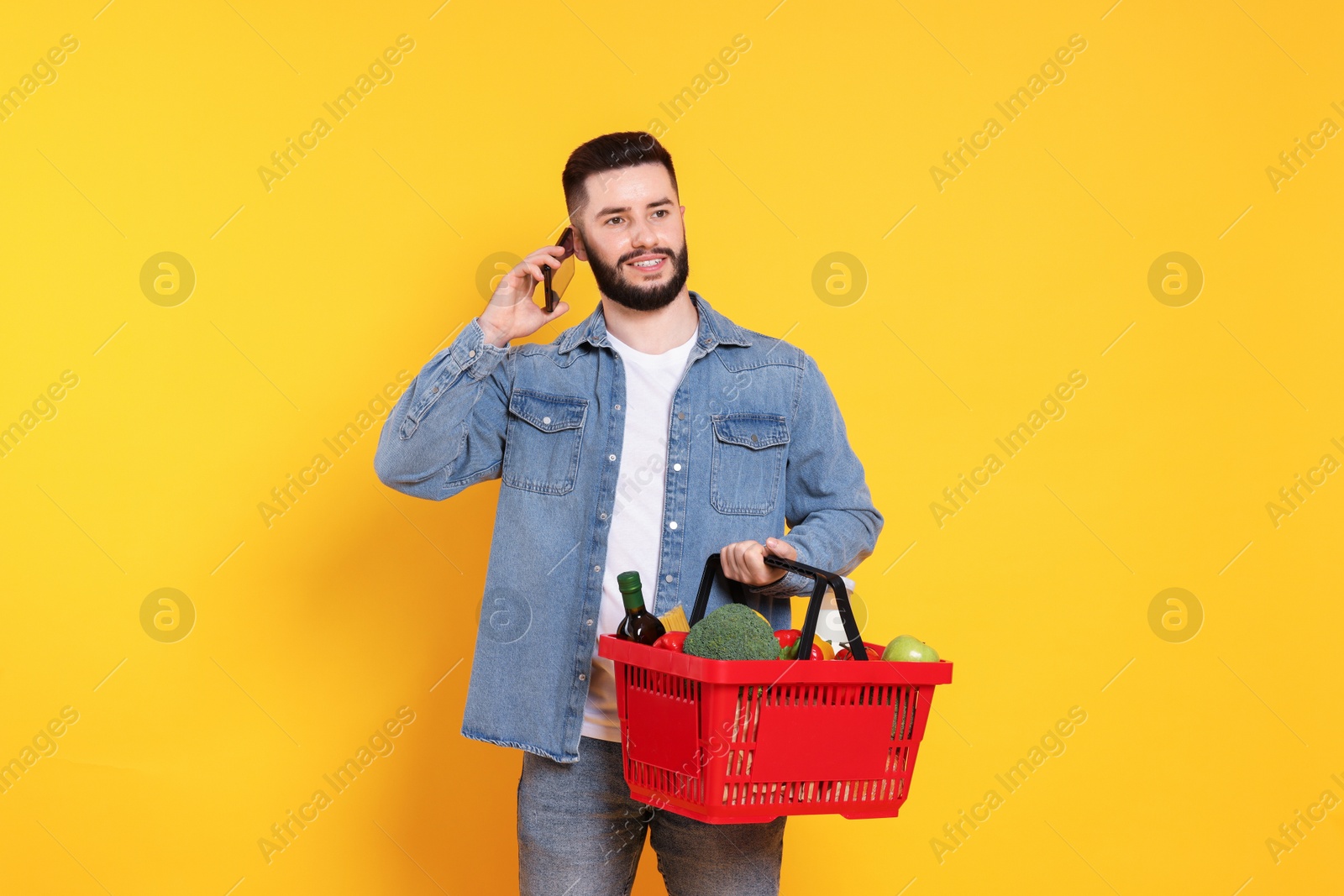 Man with shopping basket full of products talking on smartphone against orange background Photo of Man with shopping basket full of products talking on smartphone against orange background