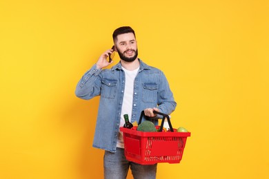 Photo of Man with shopping basket full of products talking on smartphone against orange background