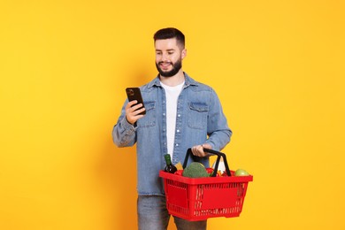 Photo of Man with shopping basket full of products and smartphone on orange background