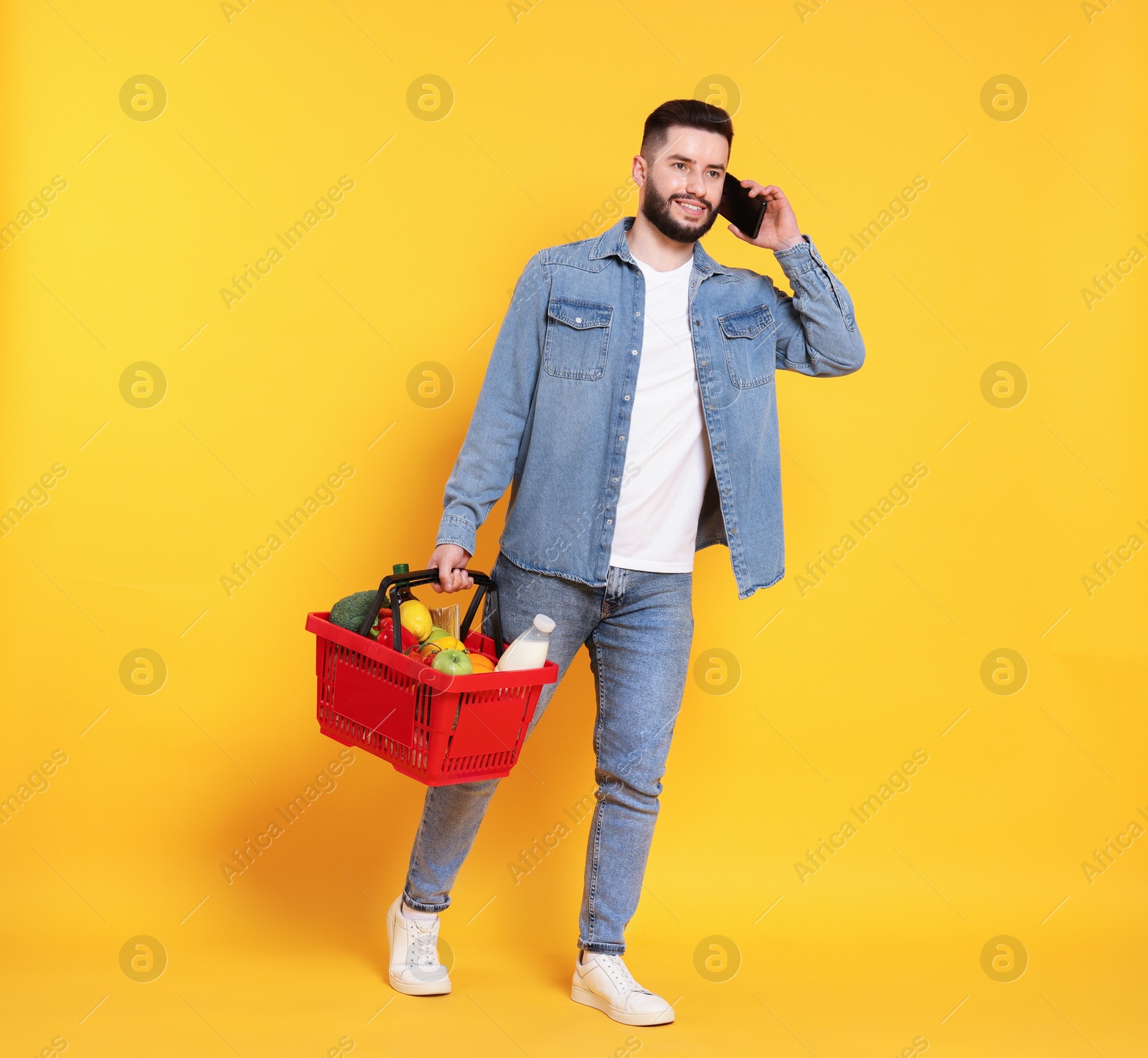 Man with shopping basket full of products talking on smartphone against orange background Photo of Man with shopping basket full of products talking on smartphone against orange background