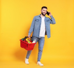 Photo of Man with shopping basket full of products talking on smartphone against orange background