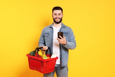 Photo of Man with shopping basket full of products and smartphone on orange background