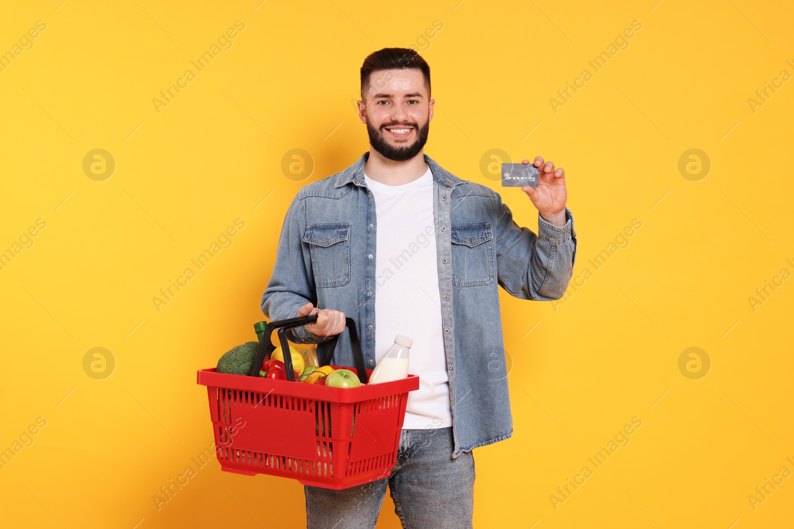 Man with shopping basket full of products and credit card on orange background Photo of Man with shopping basket full of products and credit card on orange background