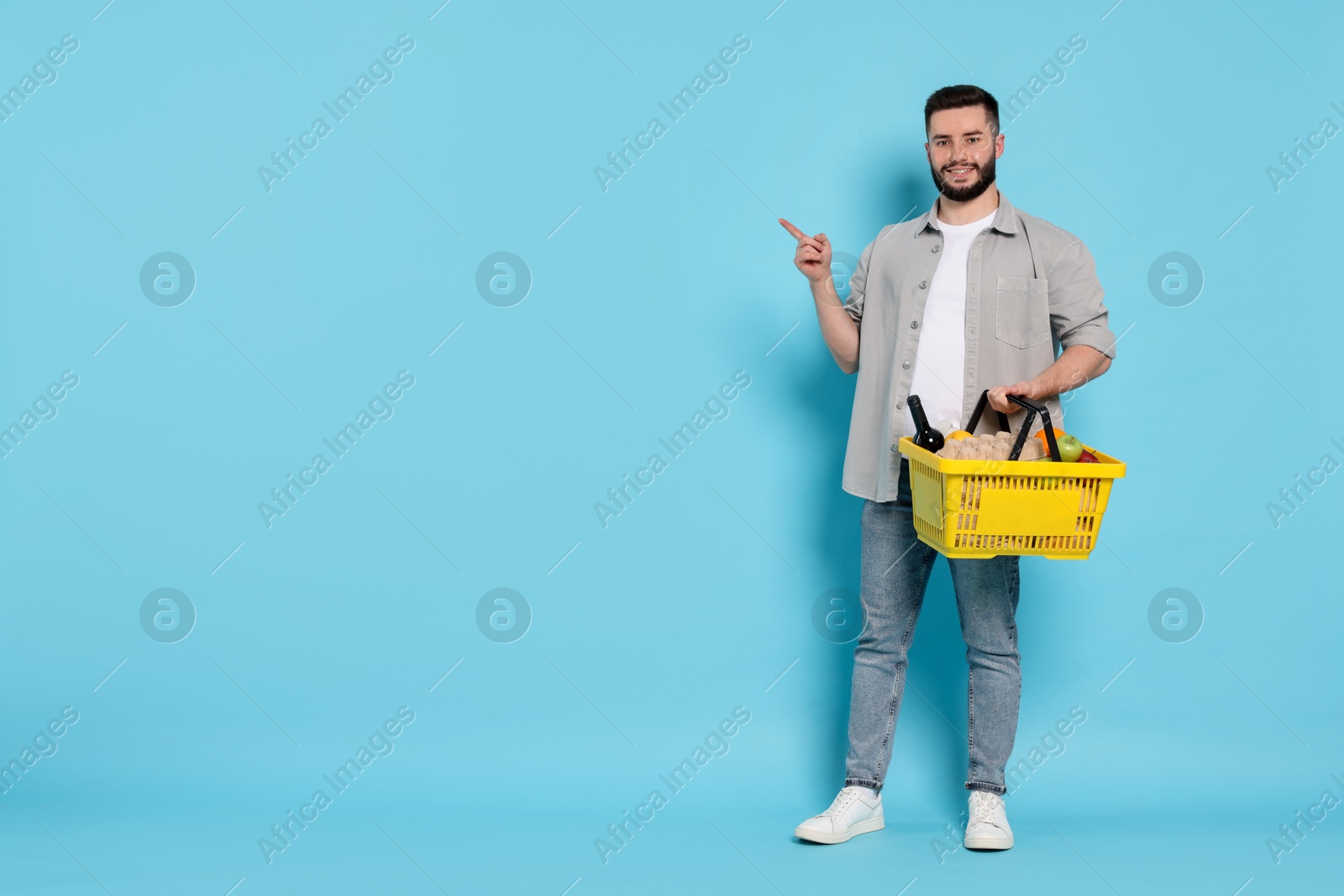 Man with shopping basket full of products pointing at something on light blue background, space for text Photo of Man with shopping basket full of products pointing at something on light blue background, space for text