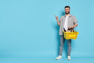 Photo of Man with shopping basket full of products pointing at something on light blue background, space for text