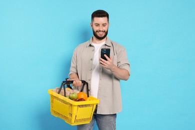 Photo of Man with shopping basket full of products and smartphone on light blue background