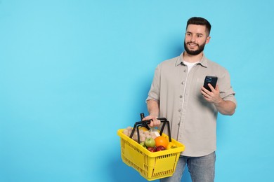 Photo of Man with shopping basket full of products and smartphone on light blue background, space for text