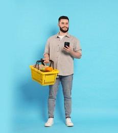 Photo of Man with shopping basket full of products and smartphone on light blue background