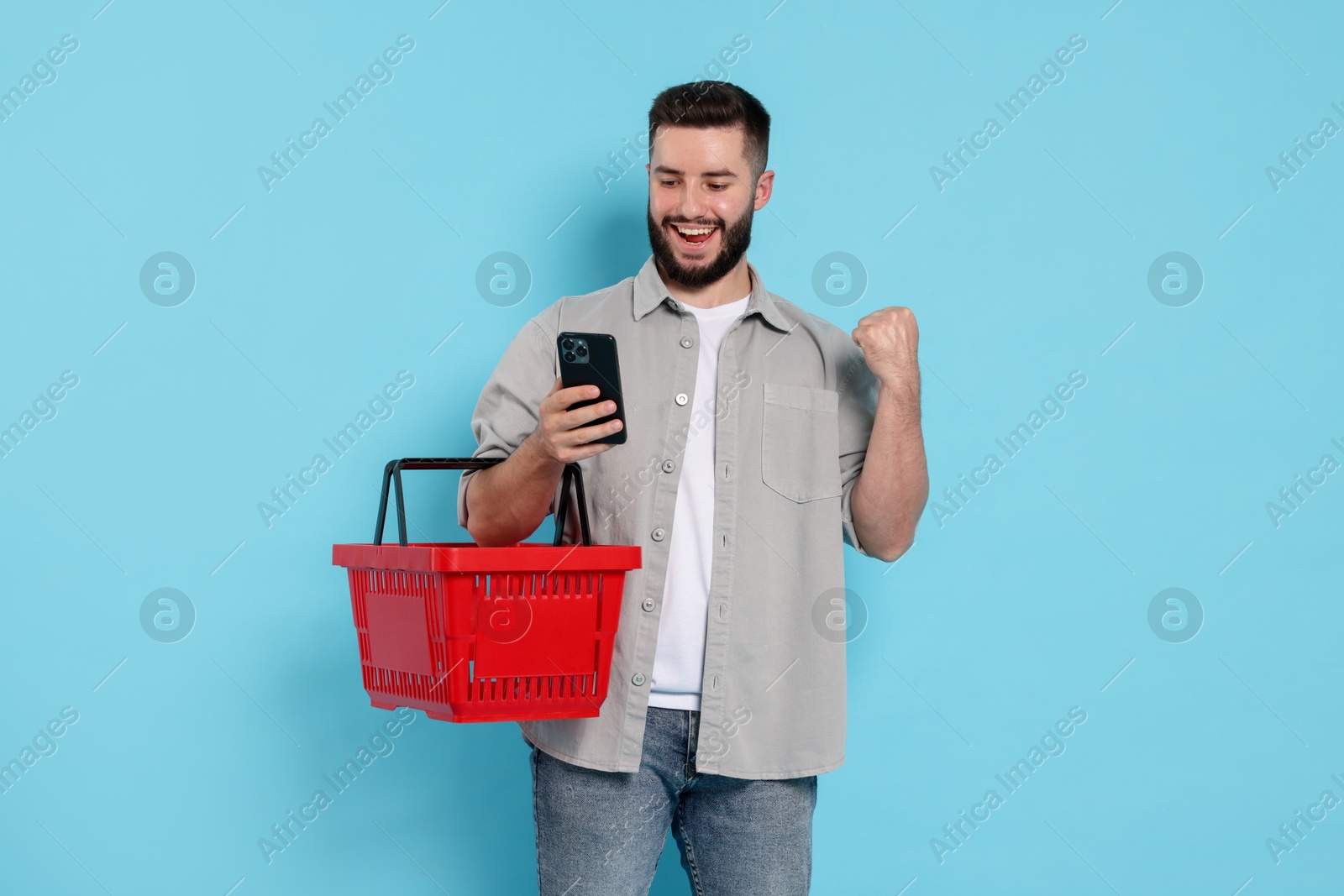 Man with empty shopping basket and smartphone on light blue background Photo of Man with empty shopping basket and smartphone on light blue background