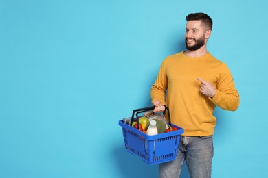 Photo of Man with shopping basket full of products pointing at something on light blue background, space for text