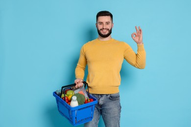 Photo of Man with shopping basket full of products showing ok gesture on light blue background