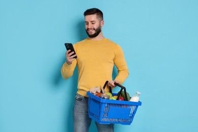 Photo of Man with shopping basket full of products and smartphone on light blue background