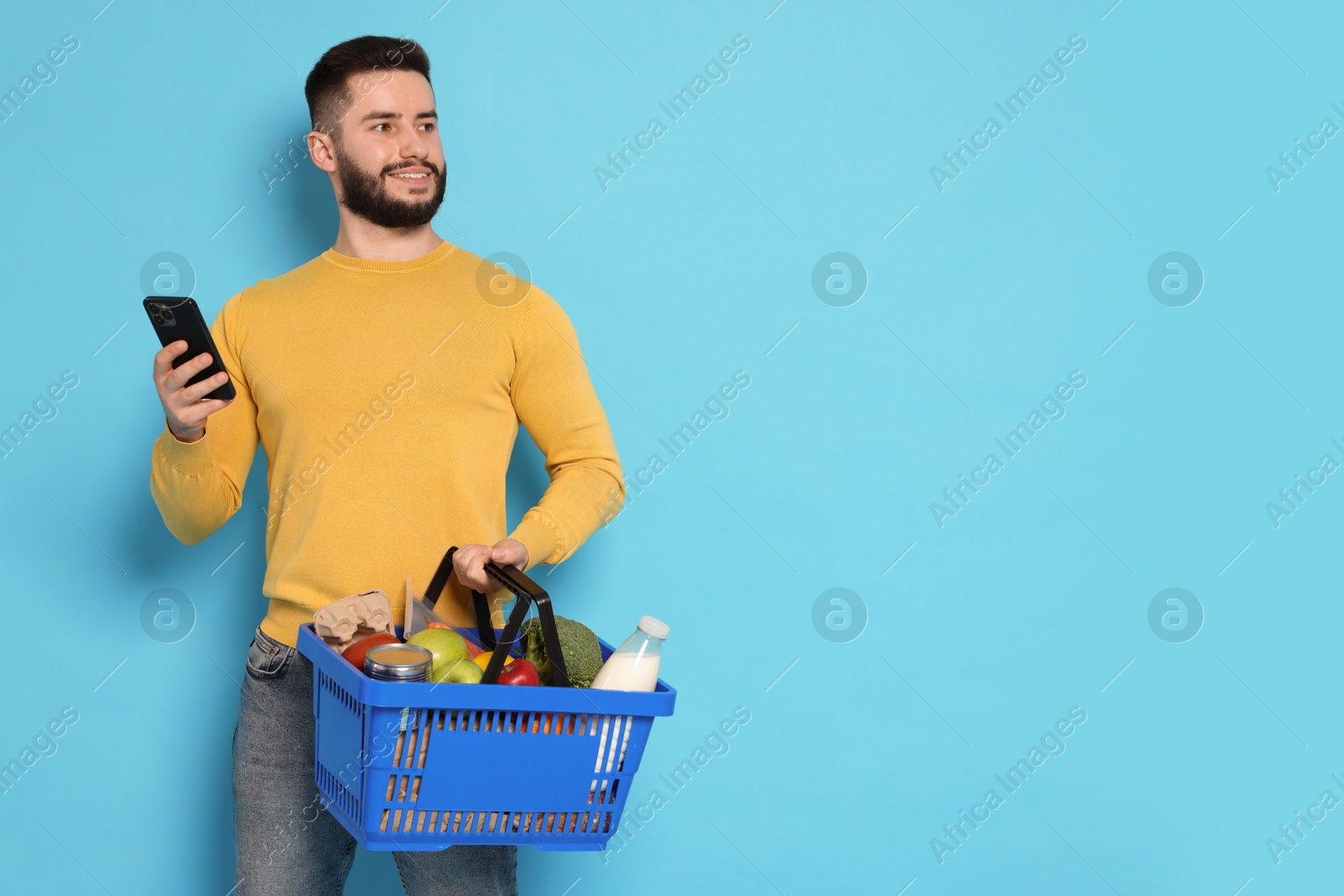 Man with shopping basket full of products and smartphone on light blue background, space for text Photo of Man with shopping basket full of products and smartphone on light blue background, space for text