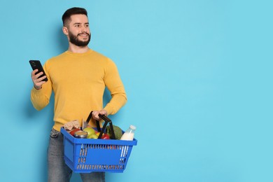 Photo of Man with shopping basket full of products and smartphone on light blue background, space for text