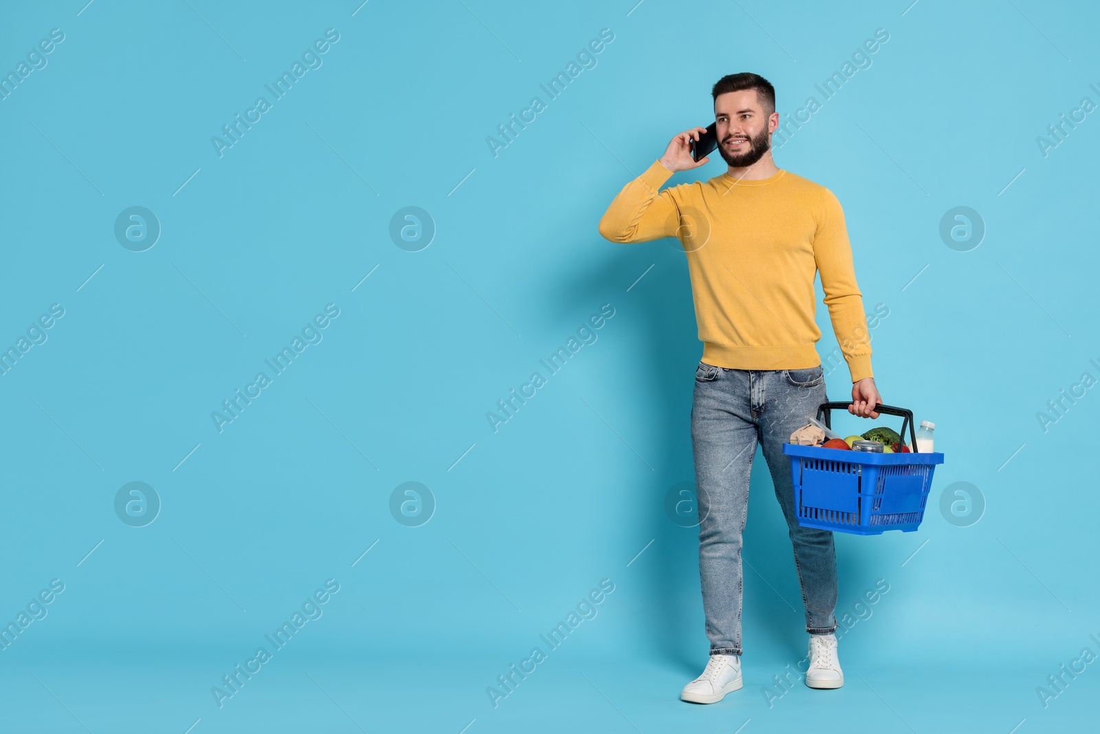 Man with shopping basket full of products talking on smartphone against light blue background, space for text Photo of Man with shopping basket full of products talking on smartphone against light blue background, space for text
