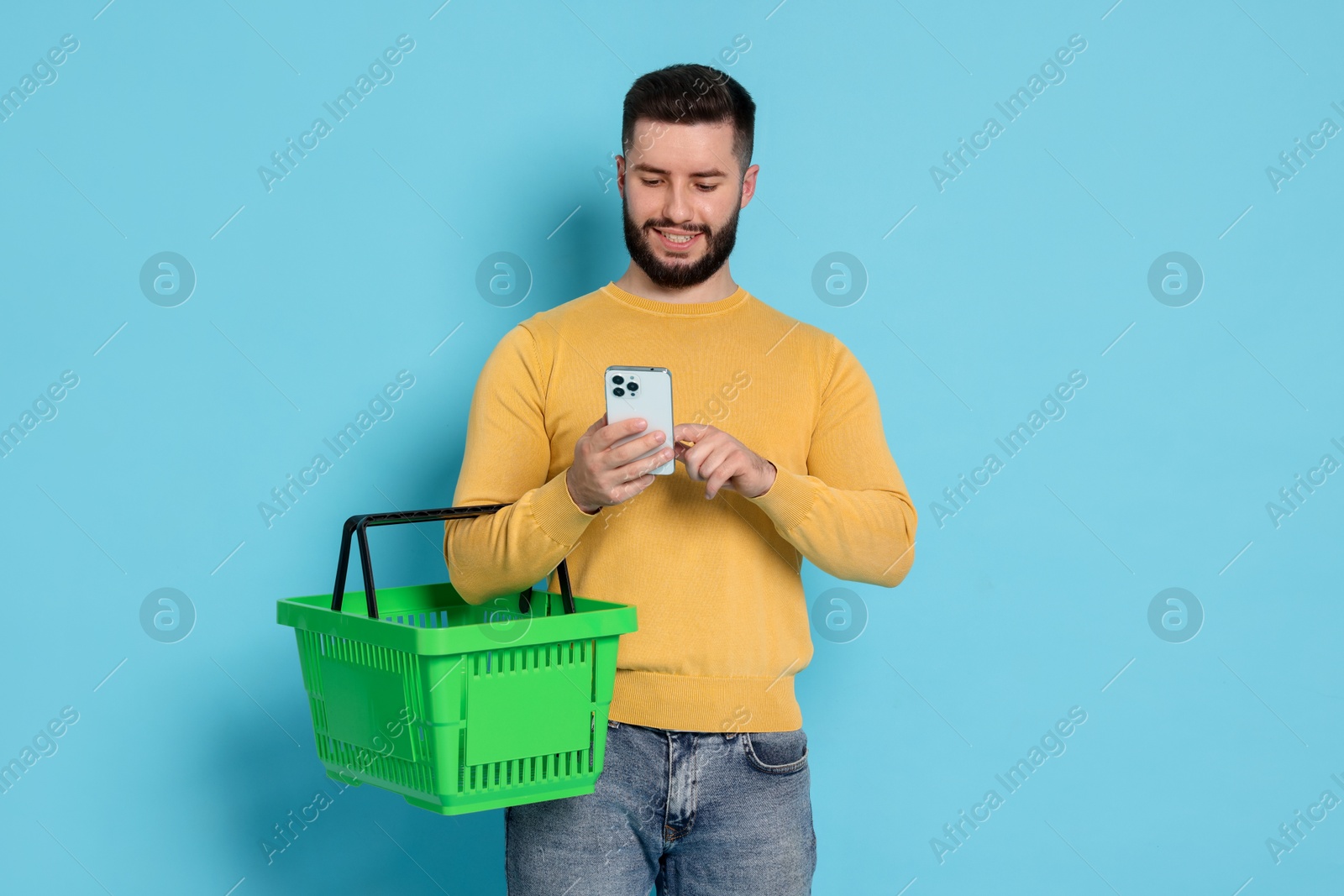 Man with empty shopping basket and smartphone on light blue background Photo of Man with empty shopping basket and smartphone on light blue background