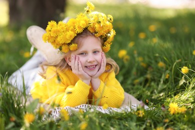 Photo of Cute little girl wearing wreath of dandelions on green grass in park, space for text
