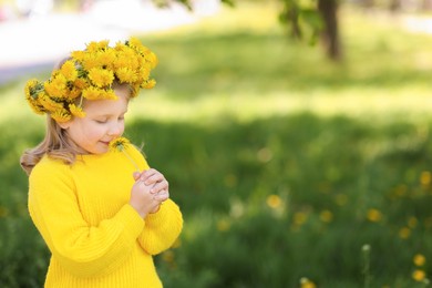 Photo of Cute little girl with beautiful flower wearing wreath of dandelions in park, space for text