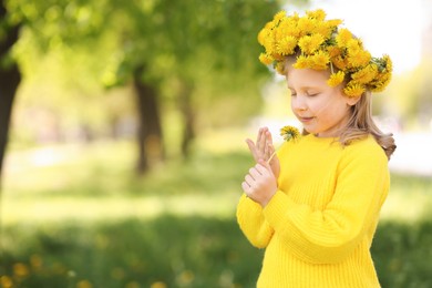 Photo of Cute little girl with beautiful flower wearing wreath of dandelions in park, space for text