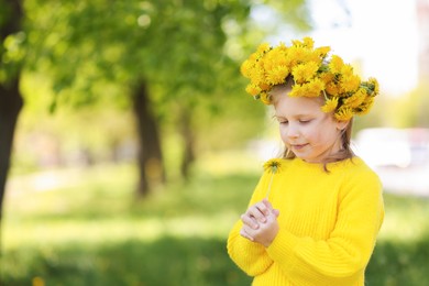 Photo of Cute little girl with beautiful flower wearing wreath of dandelions in park, space for text