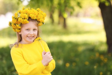 Photo of Cute little girl with beautiful flower wearing wreath of dandelions in park, space for text