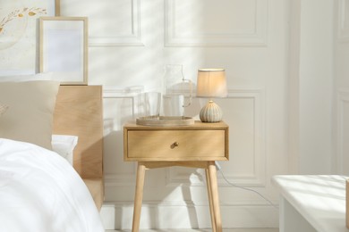 Photo of Bedside table with lamp, jug of water and glass near bed in room. Interior design